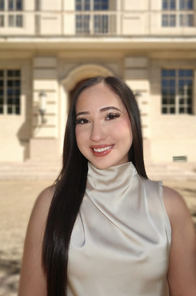 A woman with long straight dark hair, wearing a light satin sleeveless top, smiles in front of a large beige building with tall windows and an arched doorway.