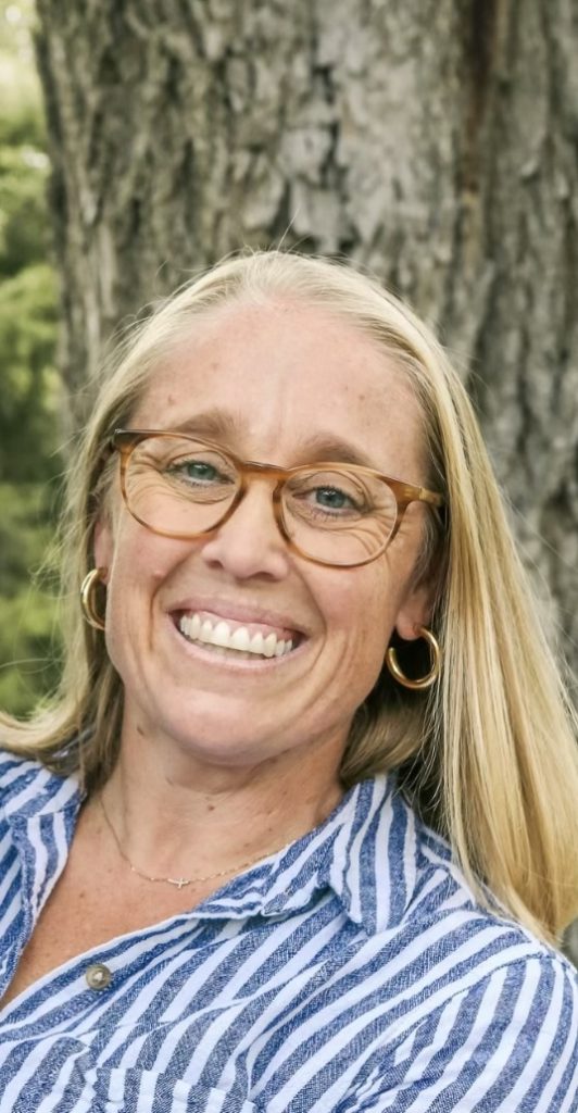 A smiling woman with long blonde hair, wearing glasses, gold hoop earrings, and a blue-and-white striped shirt, stands outdoors in front of a tree trunk.