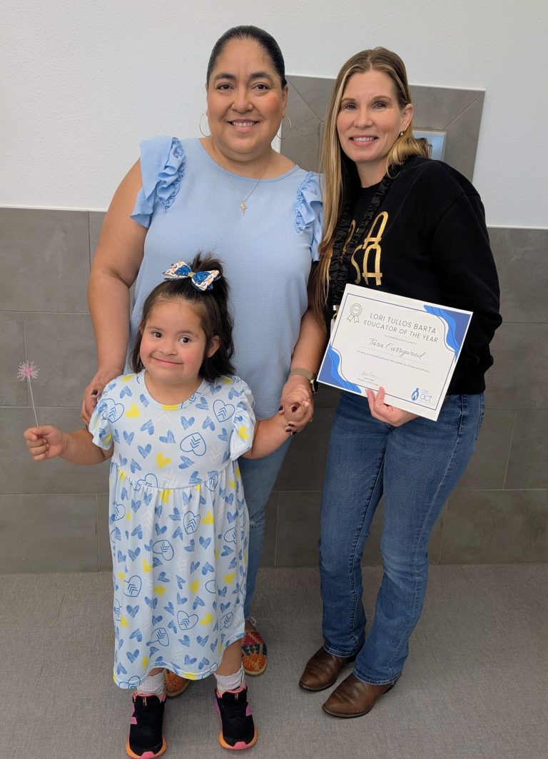 A young girl in a blue dress holds a woman’s hand and a wand, smiling. Two women stand behind her, one holding a certificate. All three are smiling indoors against a light wall and tiled floor.