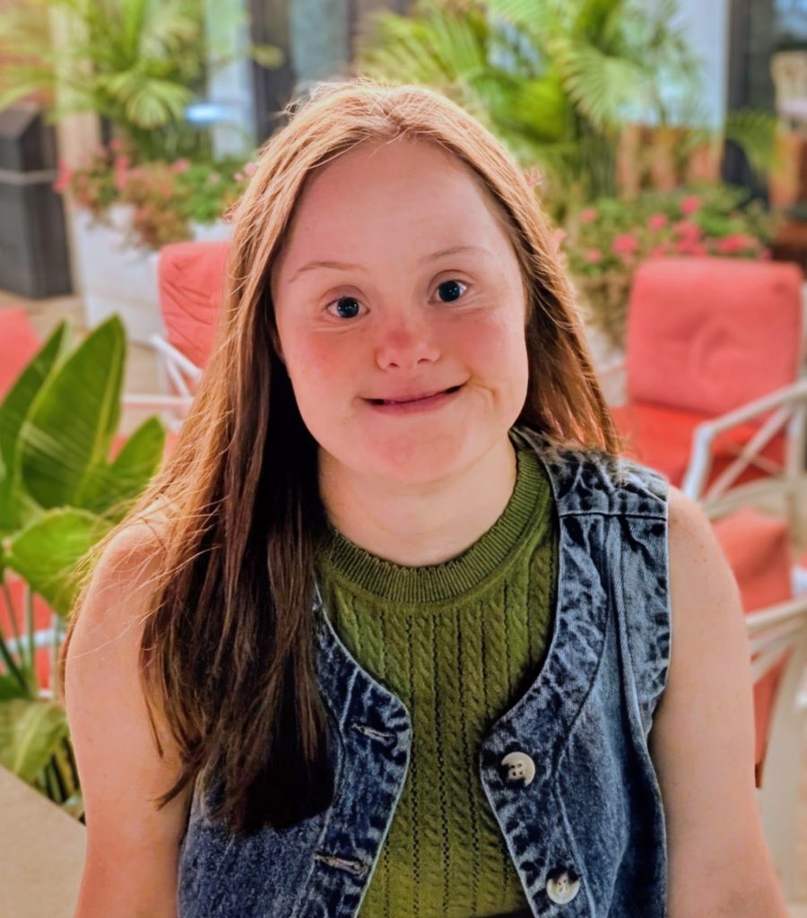 A young woman with long brown hair, wearing a green top and denim vest, smiles at the camera while sitting indoors, surrounded by plants and red-cushioned chairs.