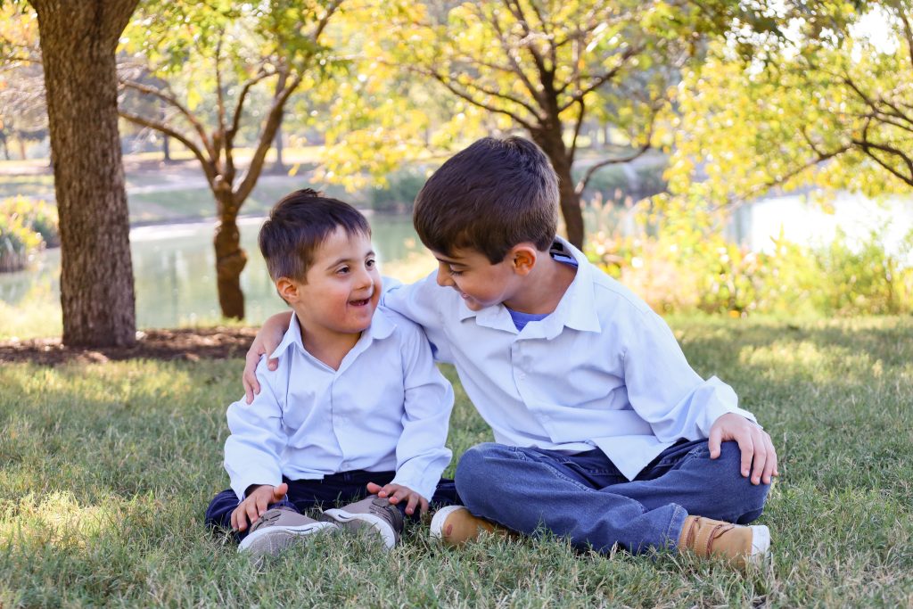 Two young boys sit on grass under trees, smiling at each other. The older boy has his arm around the younger boy. Both wear white shirts and jeans. Sunlight filters through the trees, creating a warm, cheerful atmosphere.