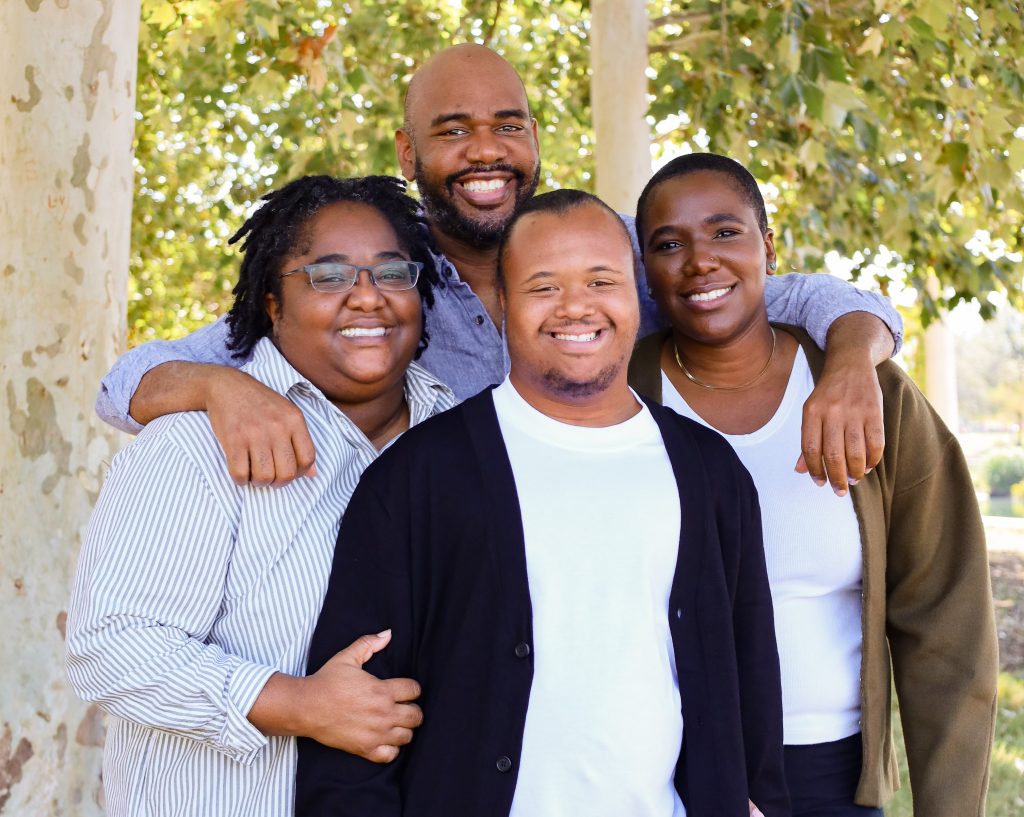 Four adults stand together outside, smiling warmly at the camera. They are in front of trees with sunlight filtering through the leaves, creating a cheerful and relaxed atmosphere.
